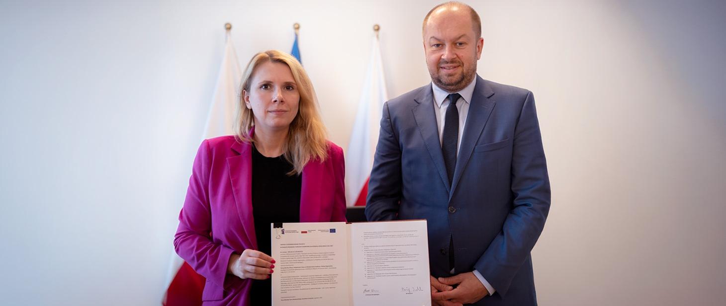 Kobieta i mężczyzna stoją obok siebie. Kobieta trzyma podpisany dokument. Za nimi widać trzy flagi.