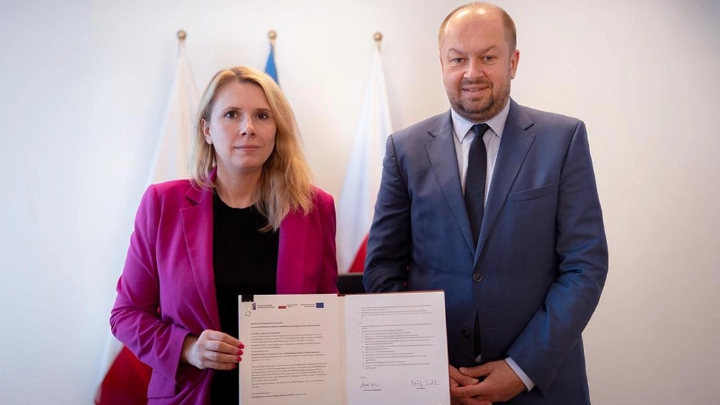 Kobieta i mężczyzna stoją obok siebie. Kobieta trzyma podpisany dokument. Za nimi widać trzy flagi.